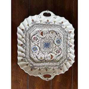 May include: A white ceramic serving platter with a scalloped edge and two handles. The center features a detailed floral and geometric design in blue, pink, and brown. The platter is approximately 12 inches in diameter.