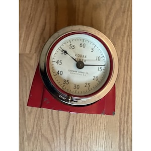 May include: A round Kodak Timer with a white face, black numerals, and black hands. The device features a chrome bezel, a red metal base, and a start-stop switch. Text on the face reads KODAK TIMER and EASTMAN KODAK CO. ROCHESTER, N.Y.