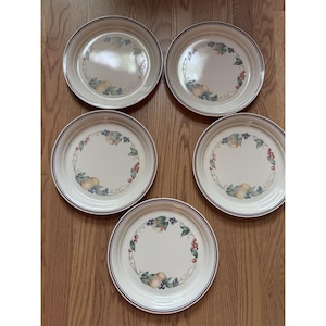 May include: Five round ceramic plates with a cream-colored base and a dark blue rim. Each plate features a central decorative wreath pattern consisting of illustrated pears, blueberries, and red cherries connected by thin, swirling brown vines.