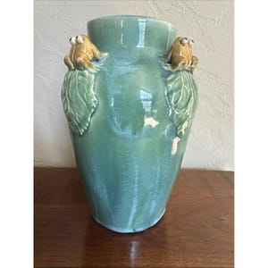 May include: A tall, turquoise ceramic vase with two frog figurines. The frogs are tan and perched on green leaf accents. The vase has a glossy finish and is displayed on a wooden surface.