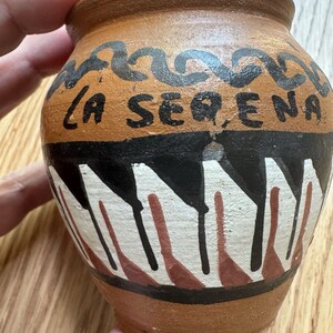 Puede incluir: Una pequeña olla de terracota pintada a mano con el texto "LA SERENA" en negro. La olla presenta diseños geométricos negros, blancos y rojos alrededor del centro y un patrón negro ondulado cerca del borde. La olla es sostenida por una mano.