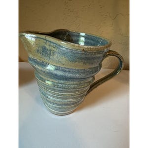 May include: A ceramic pitcher featuring a tapered, ribbed body with a glossy glaze in shades of blue, tan, and cream. The vessel includes a wide pouring spout and a curved handle attached to the side, showcasing a hand-thrown, textured surface.