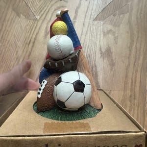 Puede incluir: Una prensa de galletas con una pelota de béisbol, fútbol americano y fútbol, con un bate y un guante de béisbol. La prensa está en una caja de plástico transparente con las palabras "cookie press" impresas.