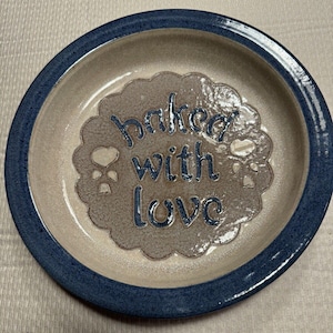 Puede incluir: Un plato de cerámica con un borde azul y un centro beige. El centro tiene un diseño decorativo con las palabras "baked with love" en azul.