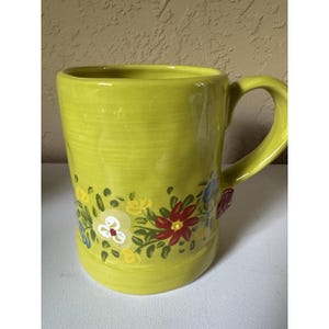 May include: A lime green ceramic mug featuring a hand-painted floral pattern around the base. The design includes red, yellow, white, and blue flowers with green leaves. The mug has a smooth, glossy finish and a curved handle on the right side.
