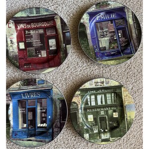 May include: A set of four round ceramic plates featuring detailed illustrations of French storefronts. The designs include a red Vins de Bourgogne bistrot, a blue Emilie shop, a blue Livres bookstore, and a green Chez Jerome restaurant facade.