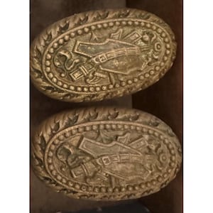 May include: Two antique gold-colored oval buttons. Each button features a detailed embossed design of a hand holding a weapon, surrounded by ornate scrollwork and a beaded border. The buttons have a vintage, aged appearance.