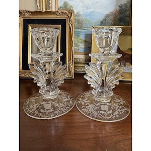 May include: Two clear glass candle holders with ornate, etched floral designs on the base. The candle holders have a detailed, fan-like design below the candle cup. The background includes framed artwork.