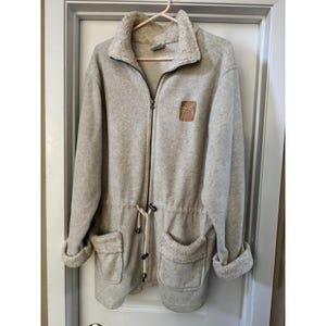 May include: A light beige fleece jacket with a full zip, drawstring waist, and two front pockets. The jacket has a beige fleece collar and cuffs. A small brown patch is sewn on the upper right chest area. The jacket is hanging on a pink hanger.