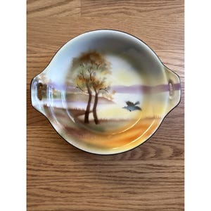 May include: A vintage, hand-painted serving dish with a landscape scene. The dish features a serene sunset with trees and a flying bird. It has two handles and a black rim, perfect for serving appetizers or small dishes.