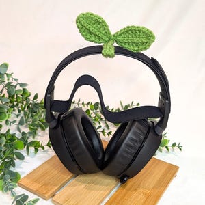 May include: Black over-ear headphones with a green crocheted leaf headband. The headphones are sitting on a wooden surface.