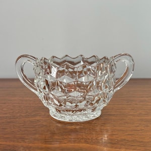 May include: A clear, vintage glass sugar bowl with a geometric pattern. The bowl has two handles and a scalloped rim. The sugar bowl sits on a round base and is displayed on a wooden surface.