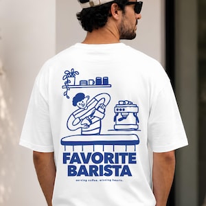 Könnte beinhalten: Ein weißes Kurzarm-T-Shirt mit einer blauen Grafik auf der Rückseite. Das Design zeigt eine Barista-Figur hinter einem Tresen mit Espressomaschine und Regal, darunter der Text "FAVORITE BARISTA" und "serving coffee, winning hearts."