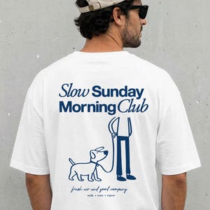 May include: A white short-sleeve t-shirt featuring a dark blue graphic on the back. The design includes the text "Slow Sunday Morning Club" above a line drawing of a person walking a dog, with the text "fresh air and good company" below.