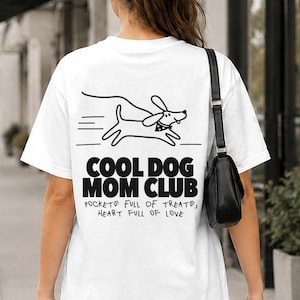 May include: A white t-shirt featuring a black line drawing of a running dog and the text COOL DOG MOM CLUB, POCKETS FULL OF TREATS, HEART FULL OF LOVE. The shirt is paired with blue jeans and a black shoulder bag, with a brown dog on a lead.