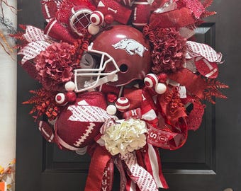 May include: A decorative wreath featuring a maroon football helmet with a white facemask, surrounded by red and white ribbons, footballs, and floral accents. The wreath is adorned with white and red decorative elements.