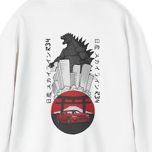 May include: White hoodie with a graphic of Godzilla above a cityscape, a red car, and Japanese text. The design is on the back. The hoodie has a drawstring hood and a kangaroo pocket. The design is in black, red, and white.
