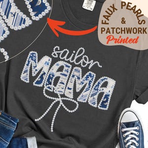 Może przedstawiać: Ciemnoszara koszulka z napisem "sailor Mama" w białych literach z imitacji pereł. Litery wypełnione są niebiesko-białym morskim wzorem patchworkowym. Widoczne są niebieskie dżinsy i niebieski but sportowy.