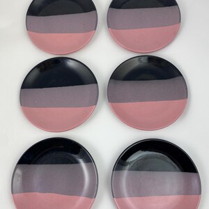 May include: Set of six ceramic plates with a three-tone design. Each plate features a black top, a gray middle section, and a pink bottom. The plates are round and appear to be handmade, with a glossy finish.