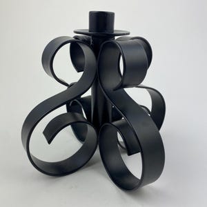Peut inclure: Bougeoir en métal noir avec un design décoratif en volutes. Le bougeoir a une colonne centrale avec une base circulaire pour la bougie et est soutenu par trois pieds en volutes.