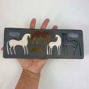 May include: Rectangular ceramic tile with a dark grey background featuring four stylized horse figures in white, dark red, and dark green. The tile is held in a hand against a white background. The horses are outlined in white, with a textured cloud design above two of the horses.