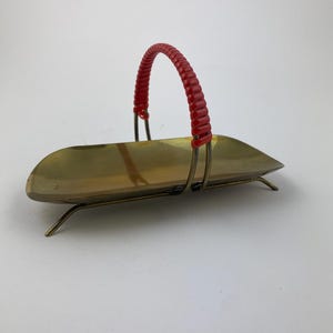 Pode incluir: Uma cesta de latão vintage com uma alça vermelha. A cesta tem uma forma oval longa com uma alça curva e suportes de arame delicados. O latão tem um tom dourado quente.