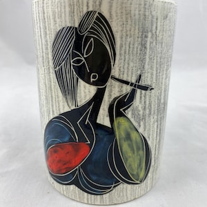 Op de afbeelding: Een cilindervormige keramische vaas met een getextureerde, gebroken witte achtergrond. De vaas heeft een zwarte en kleurrijke illustratie van een rokende vrouw. De jurk van de vrouw is blauw met rode en gele accenten. De vaas is ongeveer 13 cm hoog.