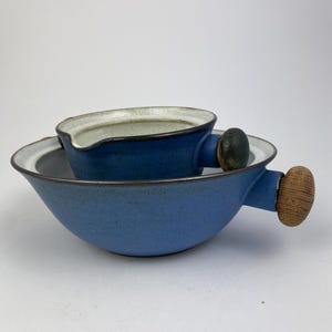 Op de afbeelding: Tweedelige blauwe keramische serveerset. De set bevat een kleine kan met een handvat en een grotere kom. Beide stukken hebben een gespikkelde textuur en een lichtgekleurde rand. De handvatten hebben een houtachtige uitstraling.