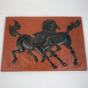 Puede incluir: Placa de pared rectangular, de color naranja rojizo, con una escultura en relieve de dos caballos encabritados. Los caballos son oscuros, posiblemente de bronce o negros, con crines y colas detalladas. La placa tiene una superficie texturizada.