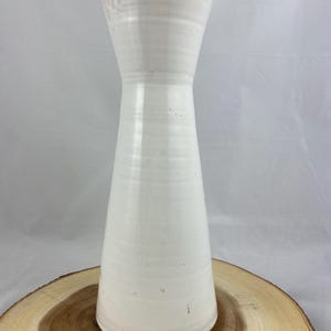 Op de afbeelding: Een hoge, crèmekleurige keramische vaas met een uitlopende bovenkant en een smalle, langwerpige body. De vaas heeft een gladde, glanzende afwerking met subtiele horizontale ribbels. De basis staat op een natuurlijke houten schijf.