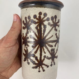 Pode incluir: Um vaso cilíndrico de cerâmica com base creme e borda marrom escuro. O vaso apresenta um padrão repetitivo de desenhos de estrelas marrons escuras. O vaso é segurado por uma mão.
