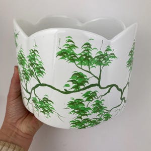 May include: White ceramic planter with a scalloped rim, hand-painted with green tree branch and leaf designs. The planter has a wide, open top and a rounded body. The design is repeated around the planter.
