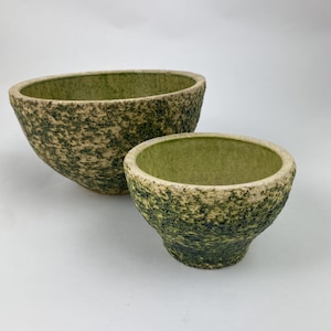 Op de afbeelding: Twee keramische kommen met textuur. De grotere kom is ovaal, de kleinere is rond. Beide hebben een gespikkelde, mosgroene buitenkant en een effen groene binnenkant. De kommen staan tegen een witte achtergrond.