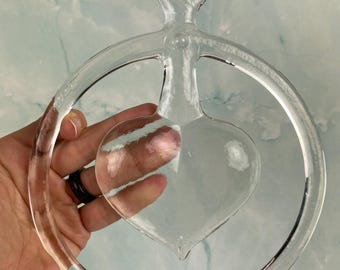 May include: A clear glass heart-shaped ornament with a circular handle and a ring. The heart is positioned within the ring. The object is held against a light blue and white background.