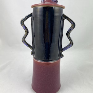 Op de afbeelding: Een zwarte en bordeauxrode keramische vaas met een deksel en twee handvatten. De handvatten zijn golvend en donkerpaars van kleur. Het deksel heeft een uniek, abstract ontwerp.
