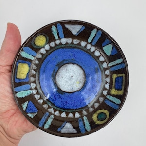 Op de afbeelding: Een klein, rond keramisch bord met een donkerbruine rand en een levendig, geometrisch ontwerp. Het midden heeft een helderblauwe cirkel met een wit centrum. De rand is versierd met blauwe, gele en witte vormen.