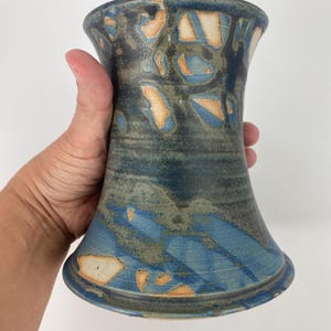 Op de afbeelding: Een handgemaakte keramische vaas met een uitlopende basis en bovenkant. De vaas heeft een blauw, grijs en oranje kleurenschema met abstracte patronen. De vaas wordt door een hand vastgehouden, wat de grootte en vorm laat zien. De vaas is ongeveer 18 cm hoog.