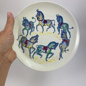 Könnte beinhalten: Ein weißer Keramikteller mit einer farbenfrohen Illustration von Zebras. Die Zebras sind in Blau, Grün, Gelb und Rot dargestellt und kreisförmig angeordnet. Der Teller hat eine leicht strukturierte Oberfläche.