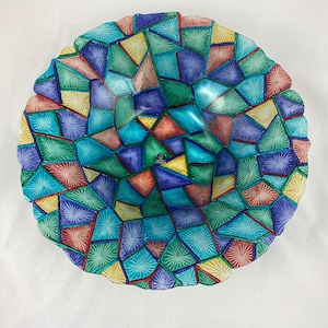 Op de afbeelding: Een decoratieve glazen kom met een mozaïekontwerp. De kom heeft een levendig patroon van geometrische vormen in tinten blauw, groen, geel en rood. De randen van de kom hebben een geschulpte vormgeving.