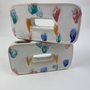 Op de afbeelding: Twee rechthoekige, witte keramische bakjes met een centrale handgreepopening. Elk bakje is versierd met kleurrijke, abstracte penseelstreken in teal, oranje, rood, blauw en groen. De bakjes zijn op elkaar gestapeld.