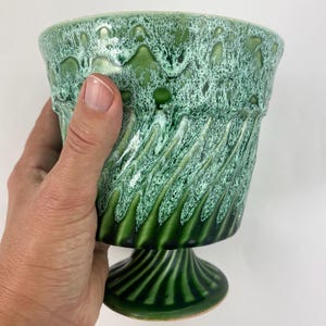 Op de afbeelding: Een groene keramische plantenbak met een voetstuk. De plantenbak heeft een getextureerd, gedruipt glazuureffect in groen- en wittinten. De voet heeft verticale groeven en de rand heeft een golvend ontwerp.