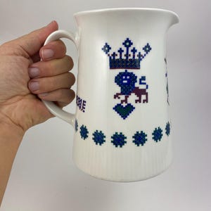 Op de afbeelding: Witte keramische kan met een handvat, versierd met een blauw en paars kroon- en leeuwenmotief. De kan heeft een rij blauwe bloemaccenten en het woord "Noble". Een traditioneel item voor het serveren van drankjes.