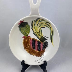 May include: A white ceramic dish with a handle, featuring a hand-painted rooster design. The rooster is depicted in brown, green, yellow, black, and red colors. The dish is on a black stand.