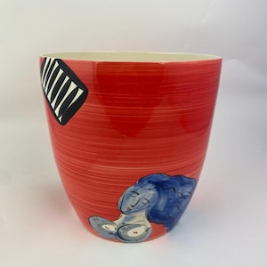Op de afbeelding: Een rode keramische plantenbak met een witte binnenkant. De buitenkant heeft een zwart-wit geometrisch ontwerp en een blauw-grijze illustratie van een liggende figuur. De plantenbak heeft een getextureerd, handgeschilderd uiterlijk.