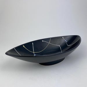 Op de afbeelding: Een zwarte keramische serveerschaal met een verhoogde voet en een langwerpige, ovale vorm. Het heeft abstracte witte, gele en blauwe lijnen. Deze schaal is geschikt voor het serveren van voedsel of als decoratief item. Ongeveer 30 cm lang.