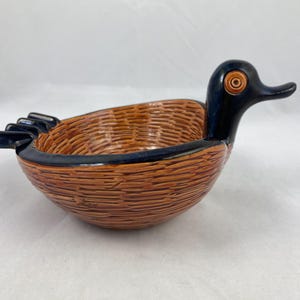 May include: A brown and black ceramic bowl shaped like a duck with a woven pattern. The duck's head is black with a brown eye.