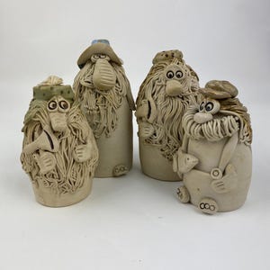 Op de afbeelding: Vier grillige keramische beeldjes met overdreven kenmerken. Elke figuur heeft een unieke hoed en baard, met expressieve ogen en neuzen. De figuren zijn in een neutraal kleurenpalet, met getextureerde details. De figuren staan op een wit oppervlak.