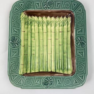 Op de afbeelding: Een rechthoekige keramische serveerschaal met een groene rand en een bruin interieur. De schaal is versierd met een rij groene asperges. De rand heeft geometrische en bladvormige ontwerpen.