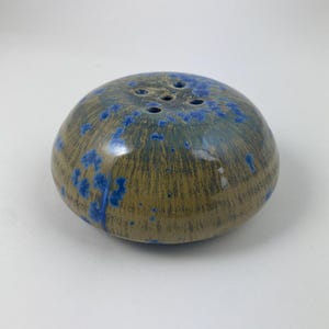 Op de afbeelding: Een keramische zoutstrooier met een afgeronde vorm en een gespikkelde glazuur. De strooier is voornamelijk bruin met blauwe accenten en heeft verschillende gaten aan de bovenkant om zout te doseren. Het ontwerp is eenvoudig en elegant.