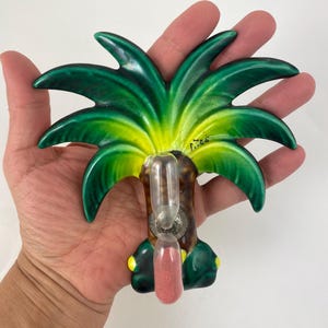 Op de afbeelding: Een keramisch object in de vorm van een palmboom met een doorzichtige glazen buis met roze zand. De palmbladeren zijn groen en geel, met een bruine stam en gele accenten. Het object wordt in een hand gehouden.